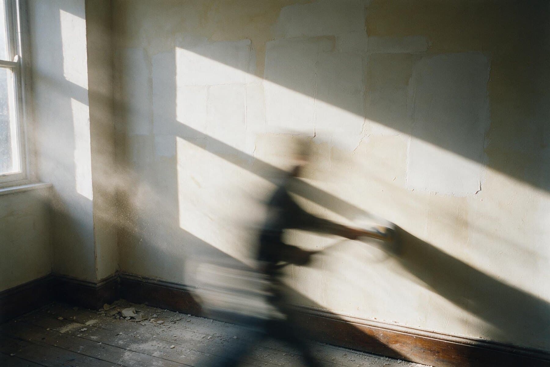 Interior wall of filled plaster patches under raking winter light, dust in the air from sanding
