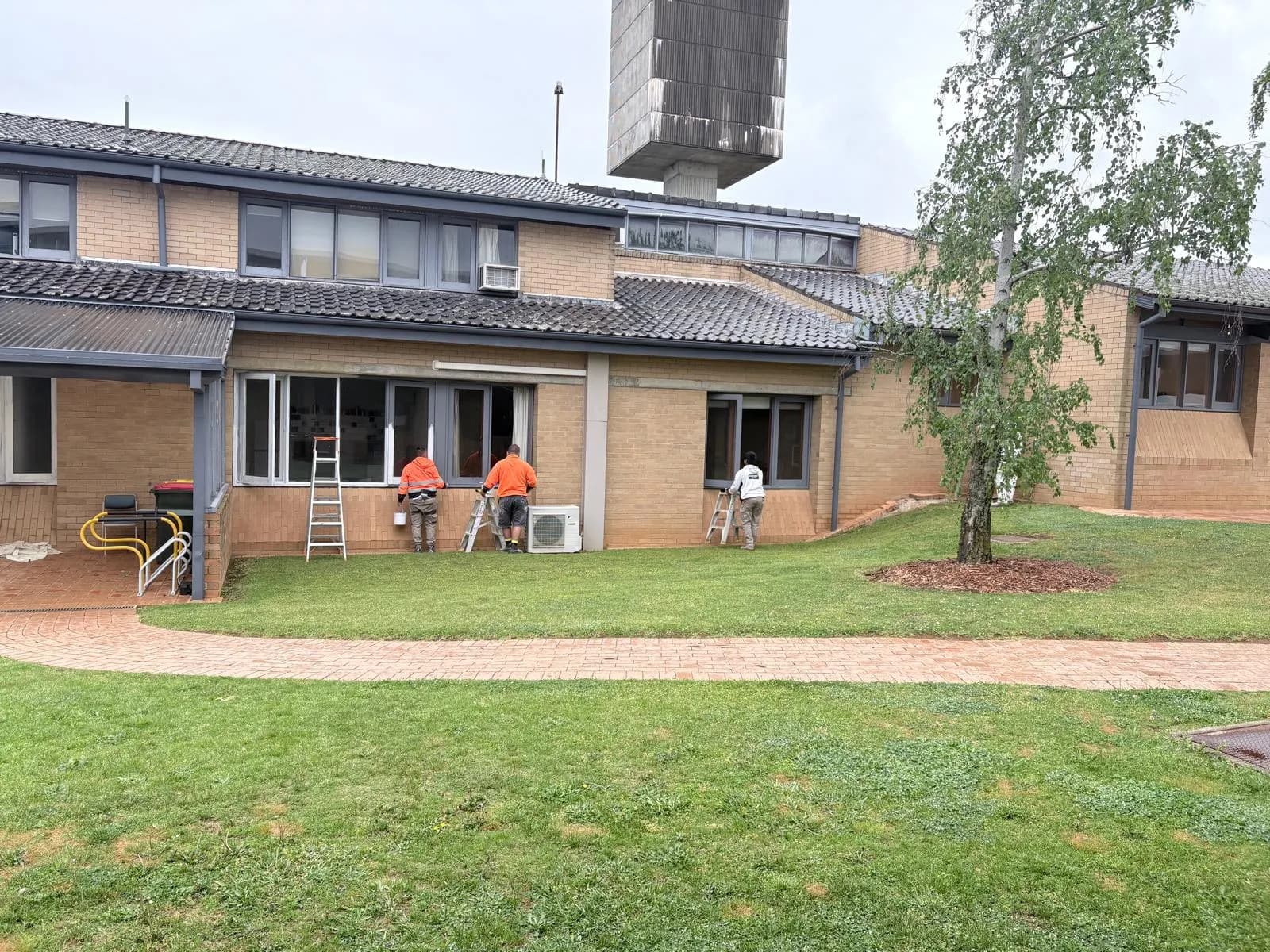 Painters on step ladders repainting window frames along a cream brick university building, in progress, Orange NSW