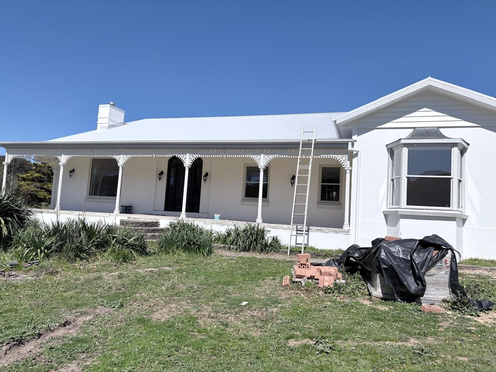 Wide view of a white rural homestead with verandah, bay window and chimney, ladder set against the front wall