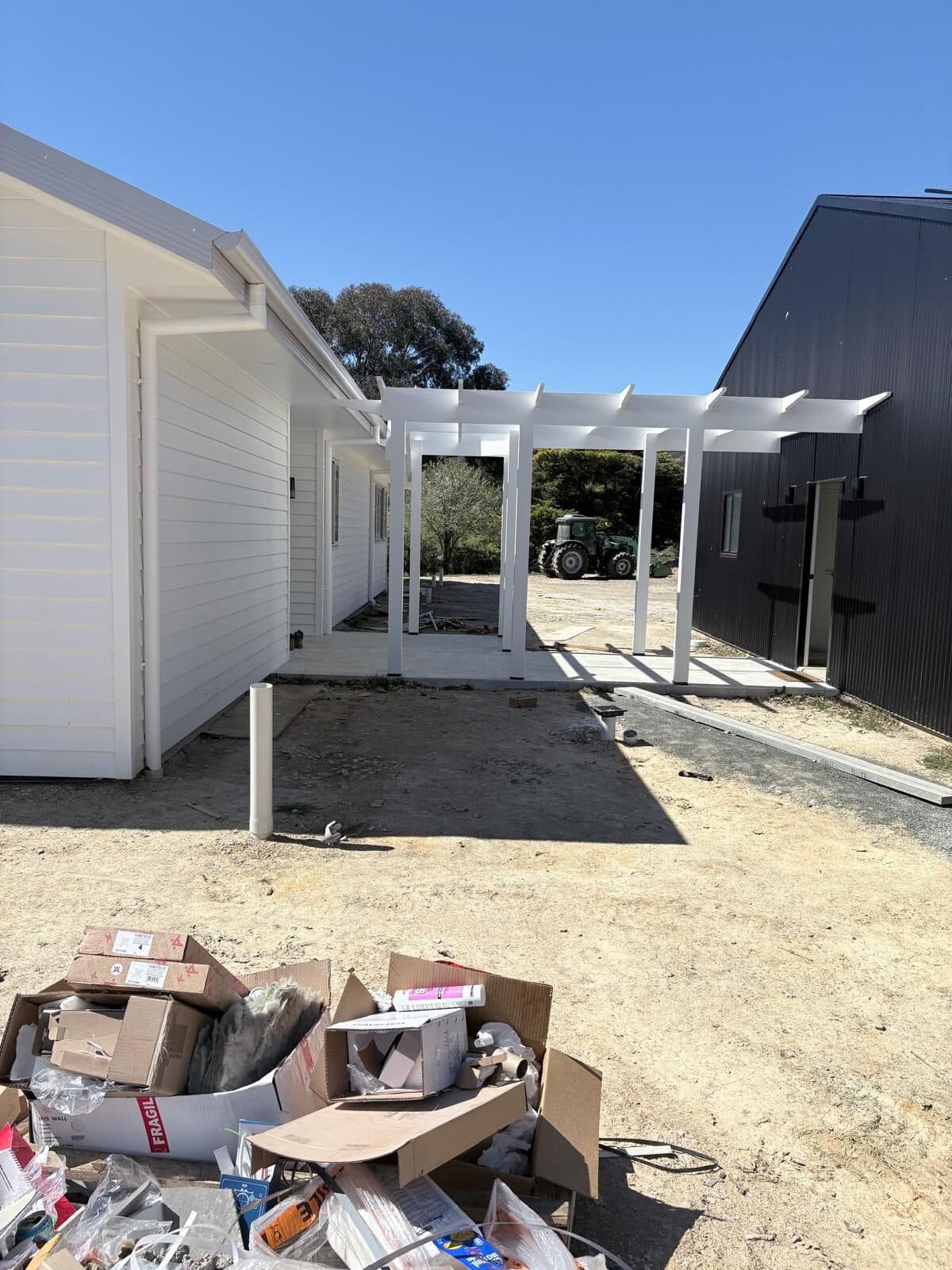 Courtyard between two wings of a rural home with a white pergola overhead, building materials still on the ground