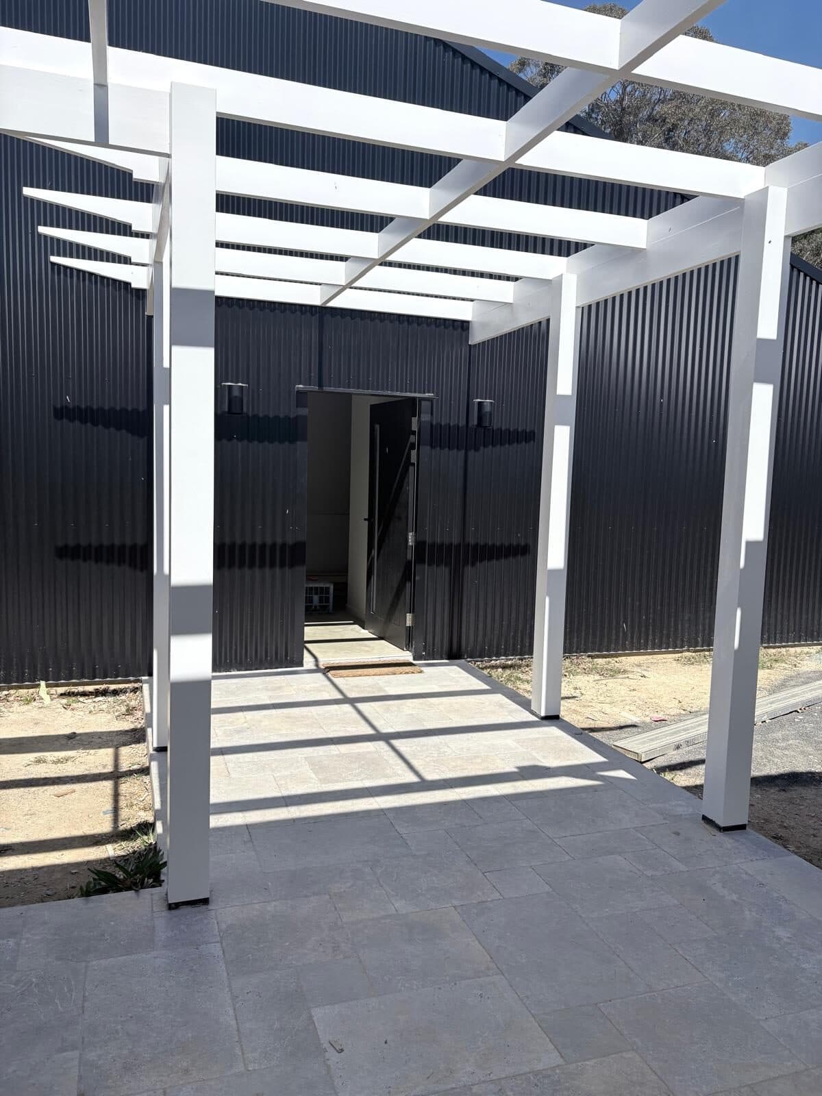 White painted timber pergola against charcoal corrugated cladding above a paved courtyard