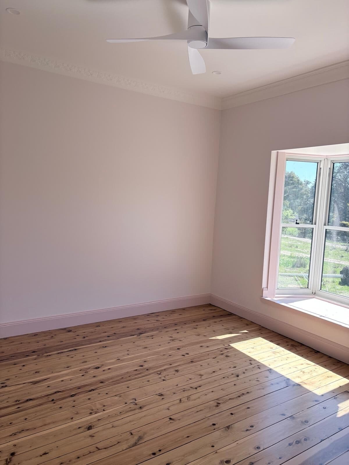 Bedroom painted in a soft neutral with a white ceiling, ceiling fan and a window looking onto rural land