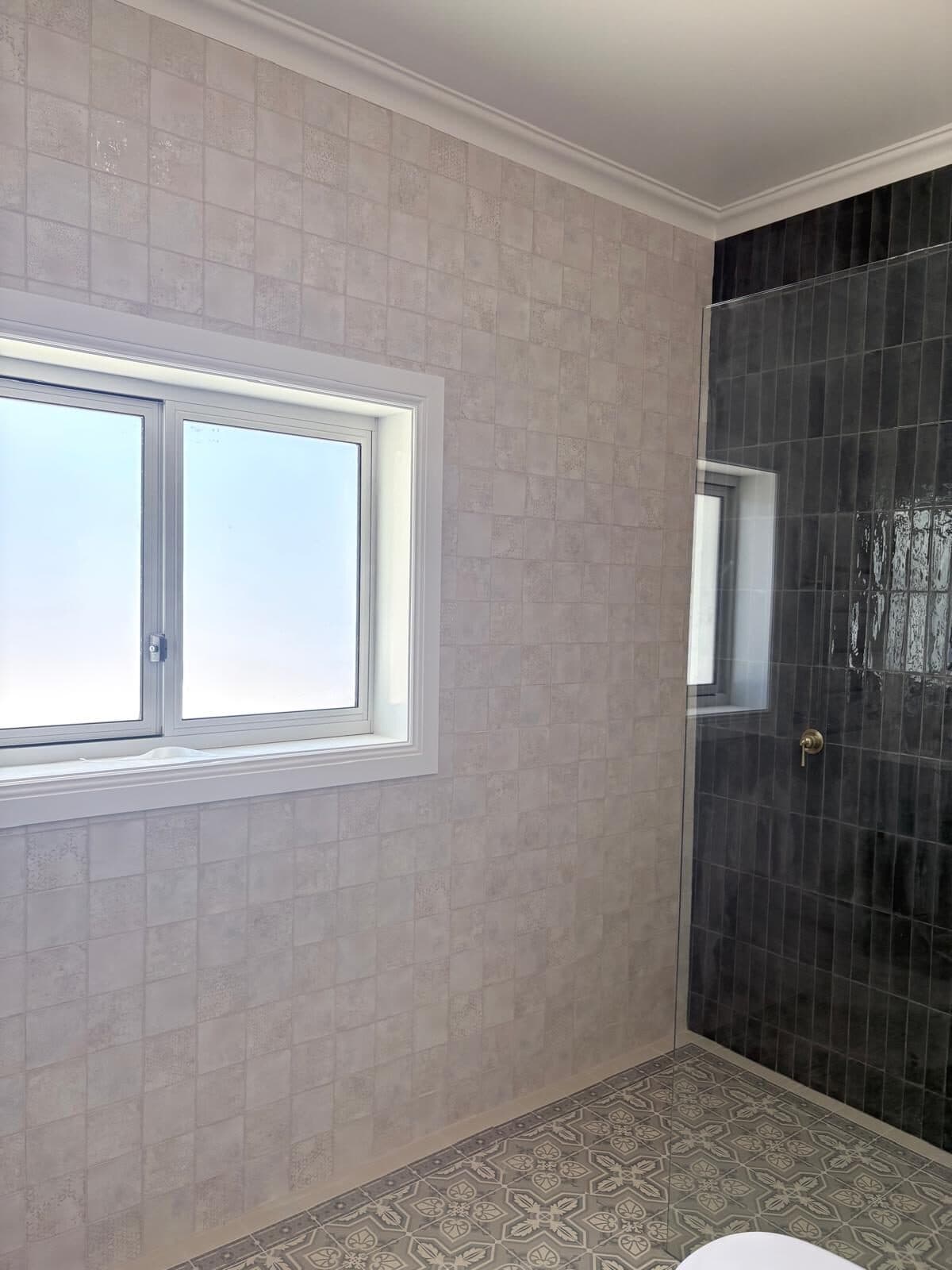 Shower recess lined in dark green glazed tile beside a window, with soft neutral walls and patterned floor tiles