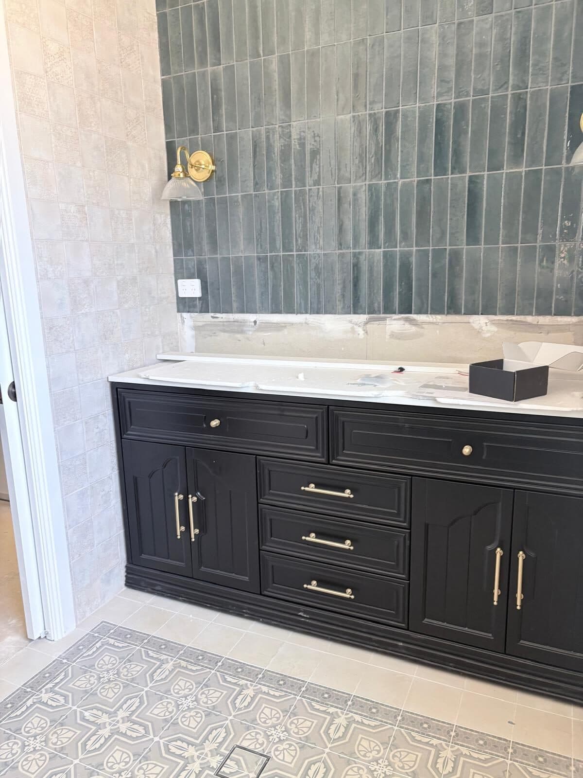 Dark painted timber vanity with brass handles below a mirror, set against green glazed tile and a patterned tiled floor