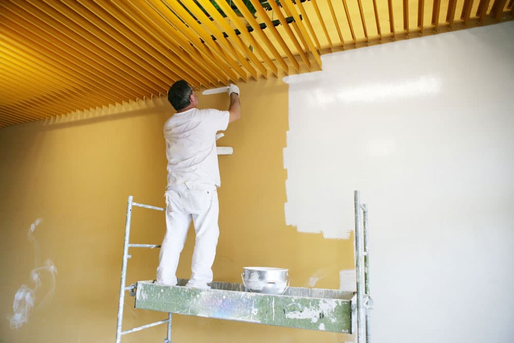 Painter in whites cutting in from a mobile trestle, paint pail on the platform and the wall part way through, Central West NSW